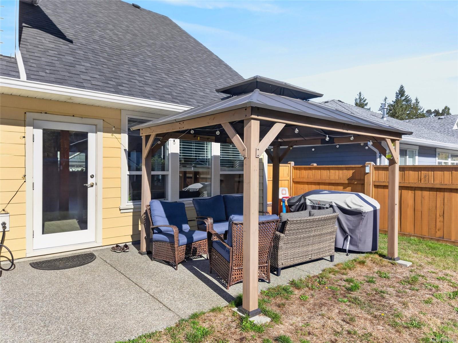 Backyard patio with a covered pergola, wicker seating, and barbecue beside a yellow-sided house.