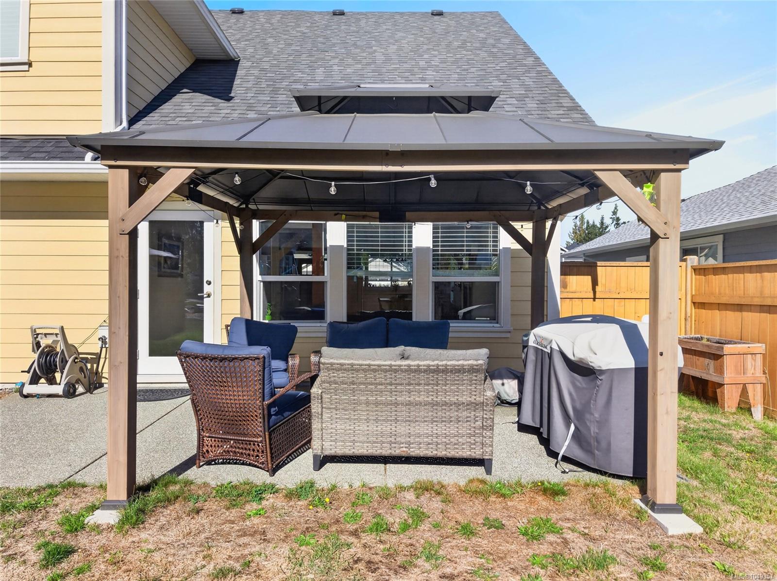 Backyard patio with a covered wooden gazebo, lounge seating, and barbecue beside a yellow-sided house.
