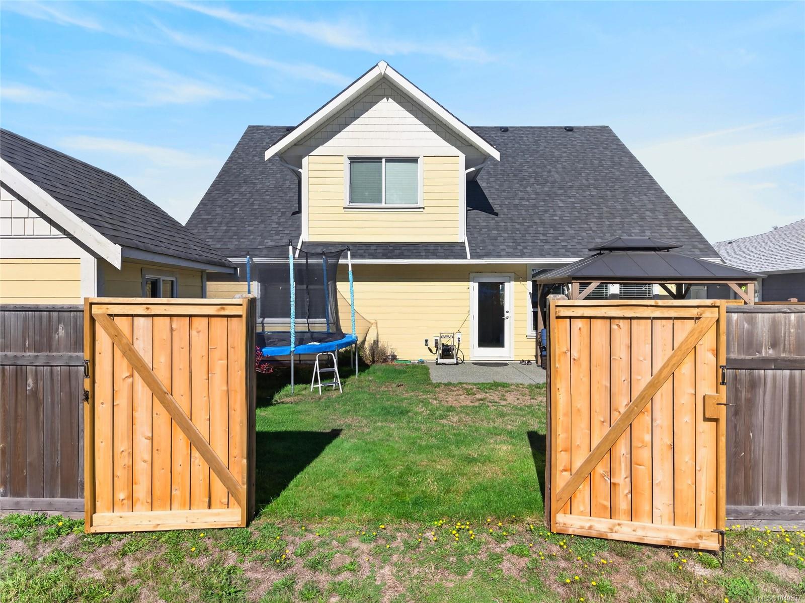 Backyard with open wooden gates, a trampoline, and a covered gazebo beside a yellow house.