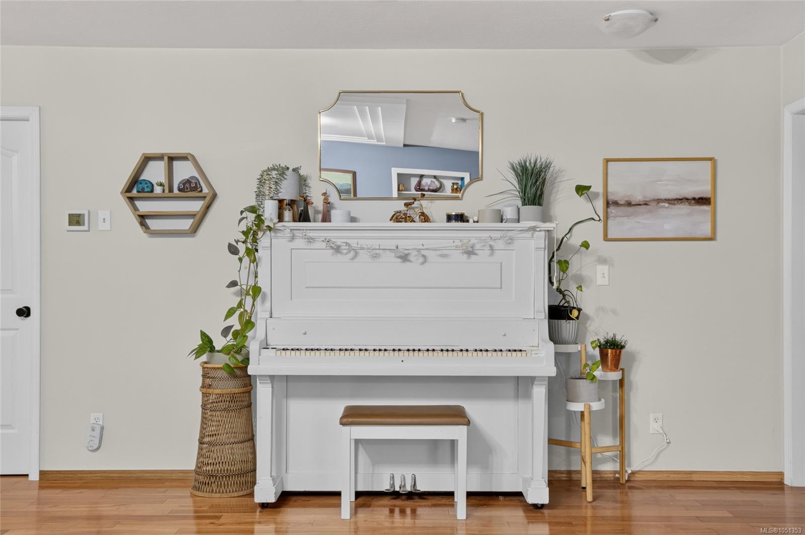 White upright piano and bench in a bright room with hardwood floors, plants, and wall decor.