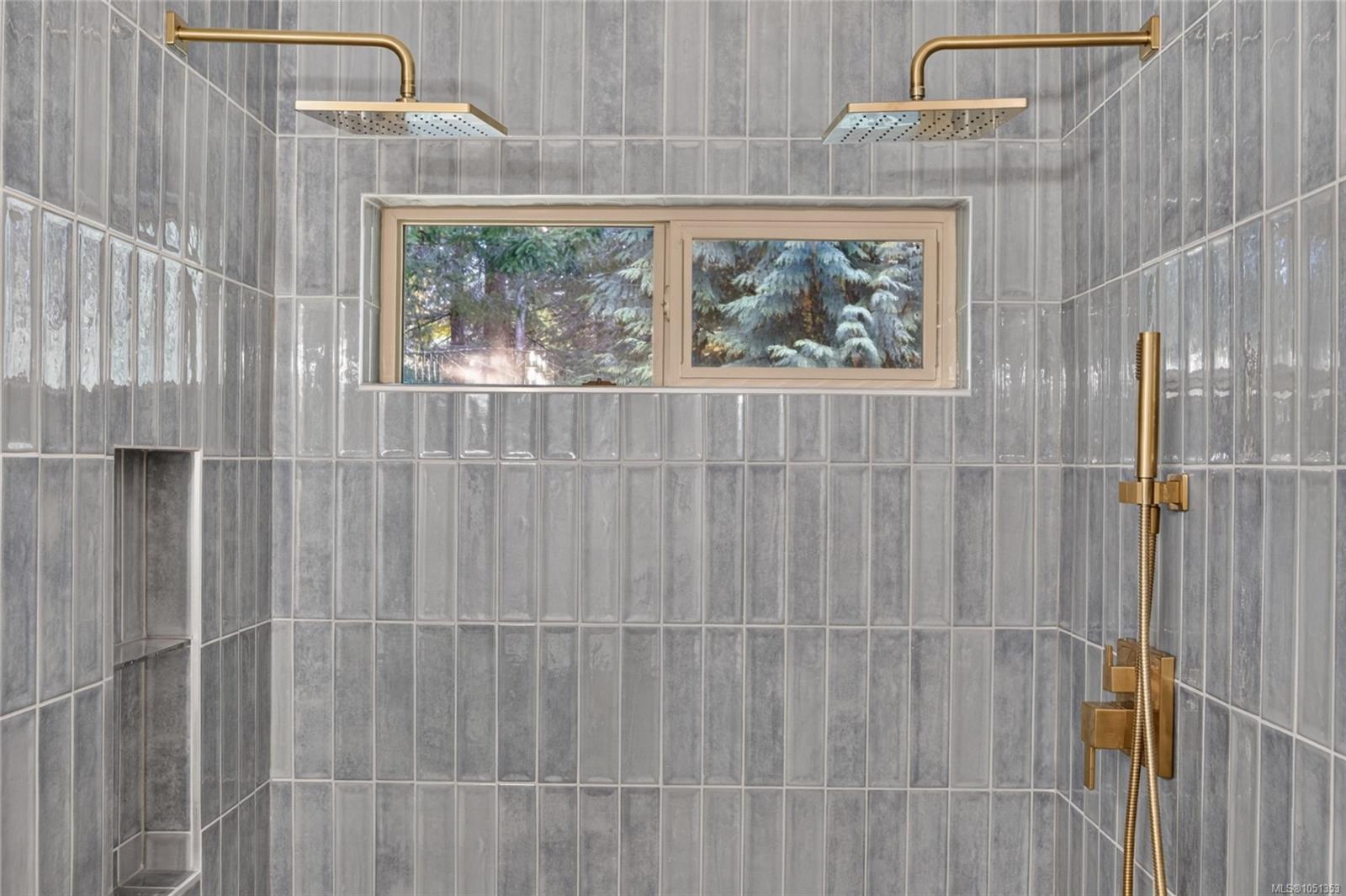 Walk-in shower with gray tile, dual brass rainfall showerheads, built-in niches, and a window overlooking trees.