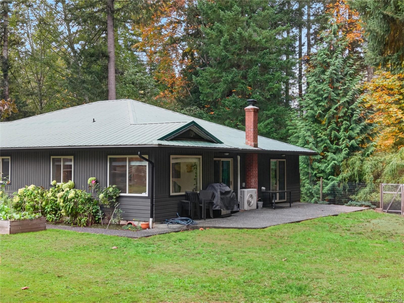 Single-story home with a pale metal roof, brick chimney, paved patio, and wooded autumn setting.