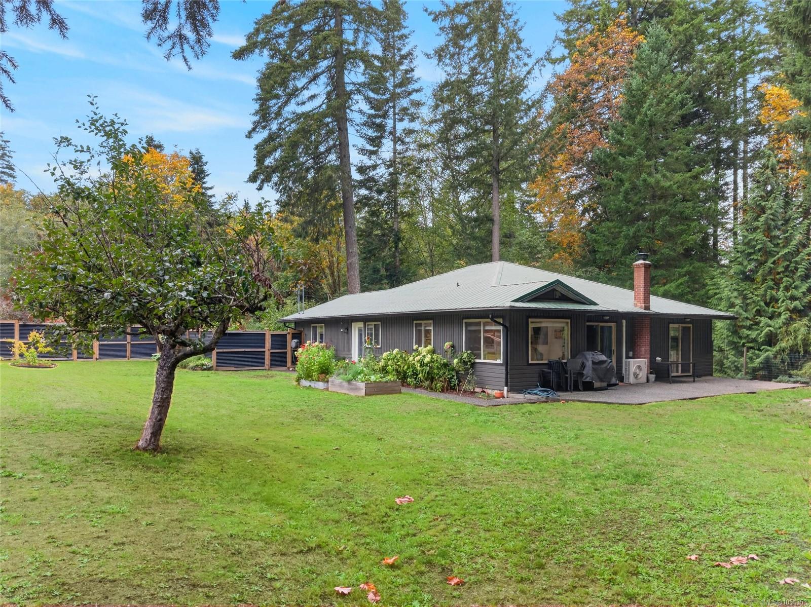 Single-storey dark-sided house with a metal roof, brick chimney, garden beds, and lawn beneath tall trees.
