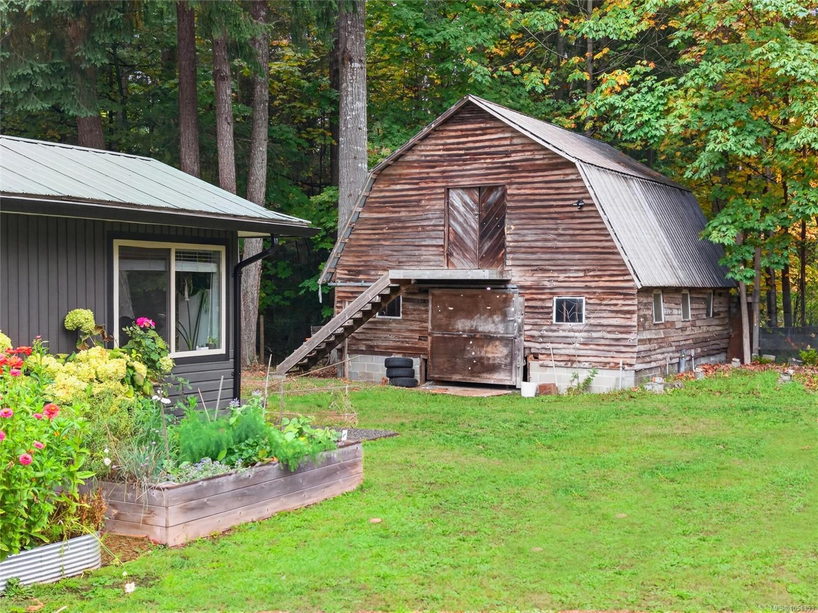 Wooden gambrel-roof barn beside a house, with garden beds and mature autumn trees.