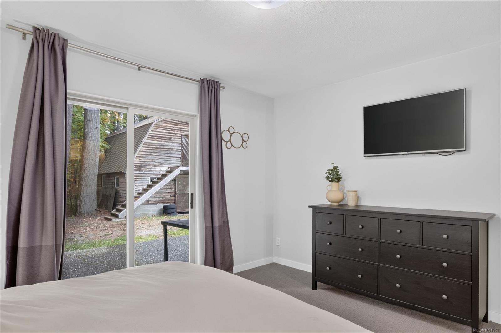 Furnished bedroom with sliding glass doors overlooking a wooded yard and rustic outbuilding.