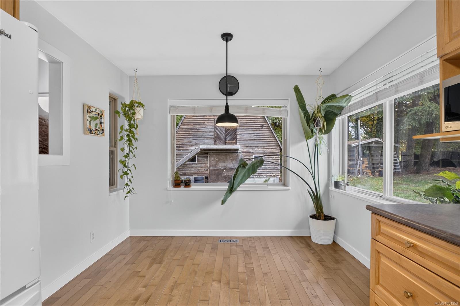 Bright kitchen with hardwood floors, wood cabinetry, and large windows overlooking a leafy yard.