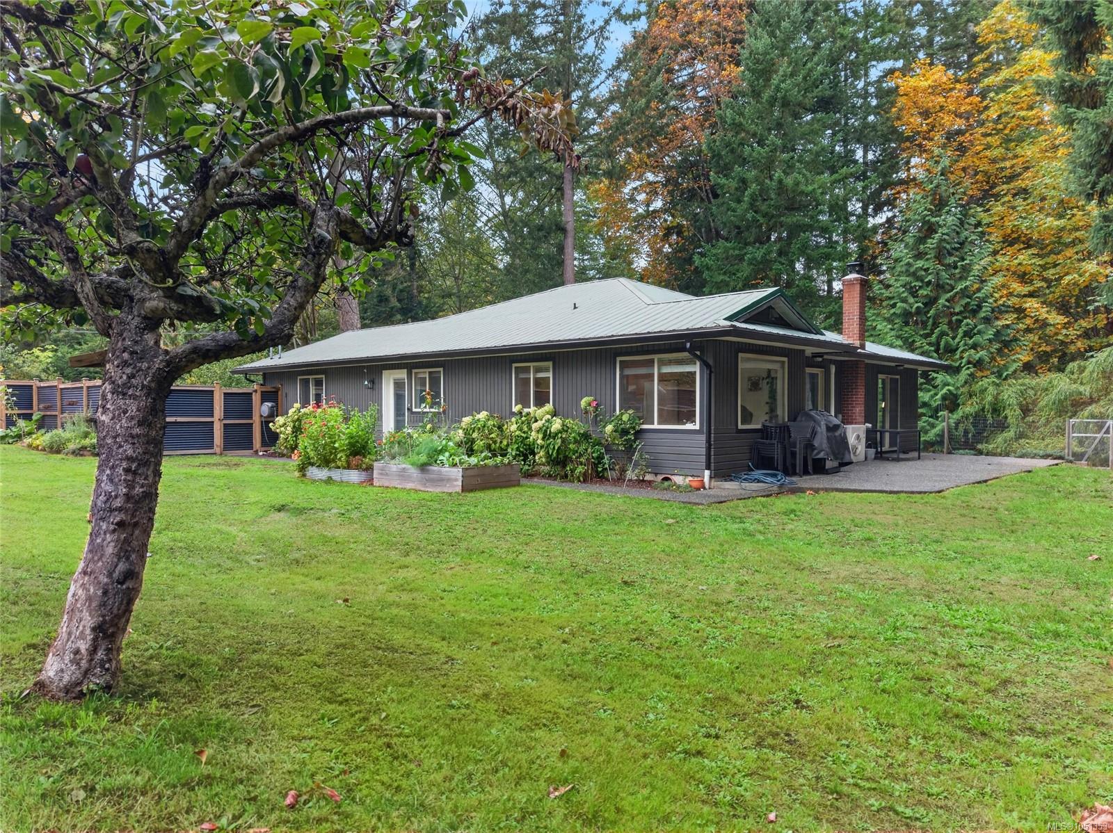 Single-story gray home with a covered patio, raised garden beds, and a wooded yard.