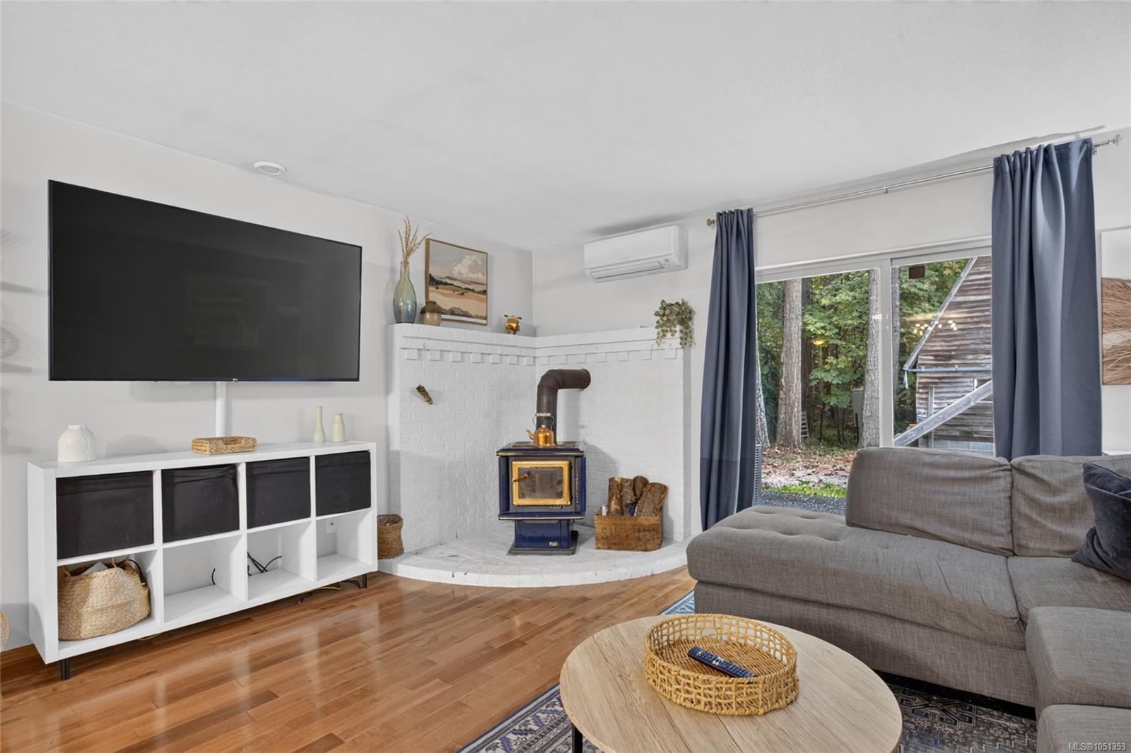 Living room with a blue wood stove, white brick fireplace, gray sectional, and windows overlooking trees.