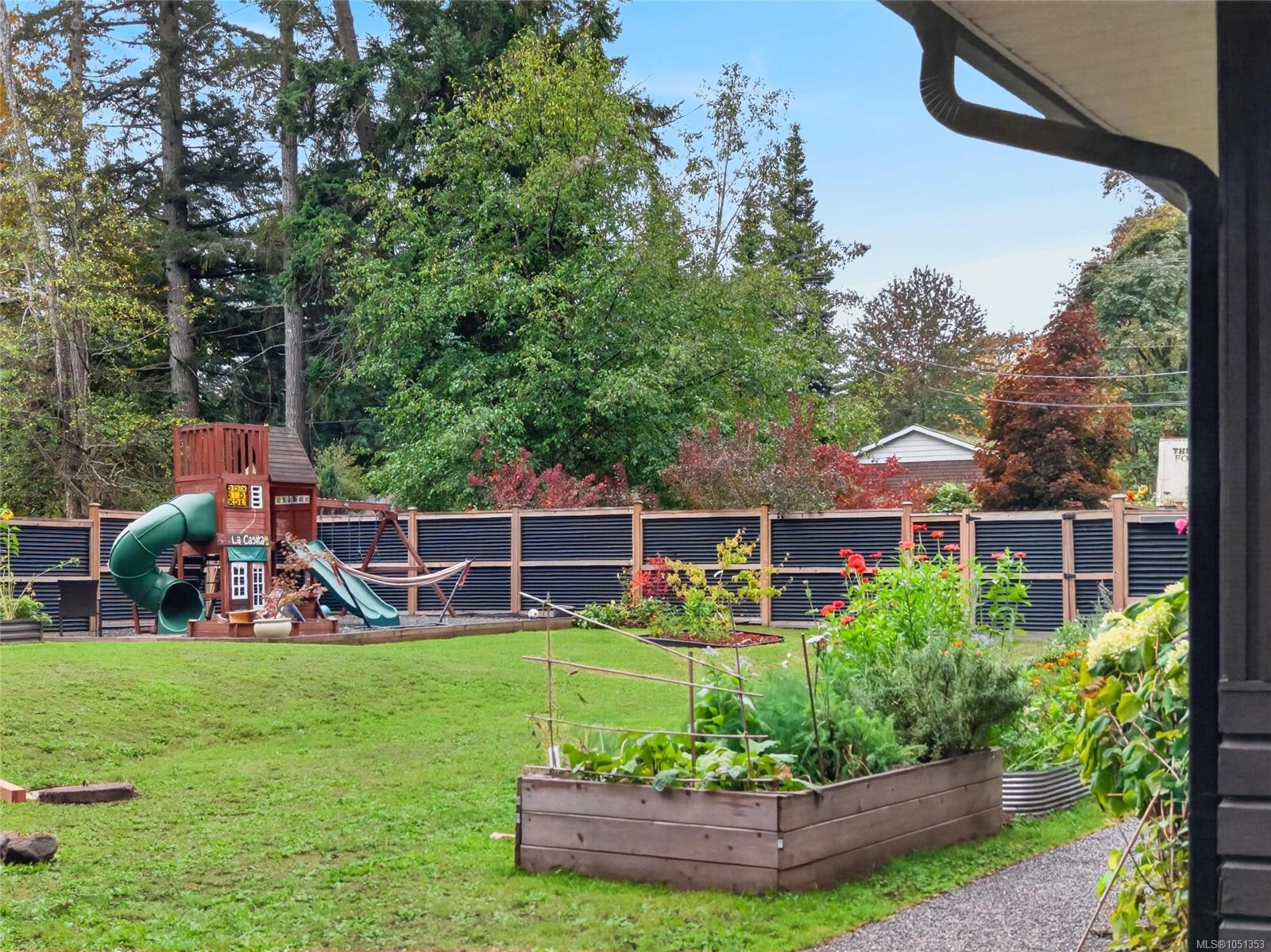 Backyard with a children’s play structure, raised garden beds, lawn, and tall fence.