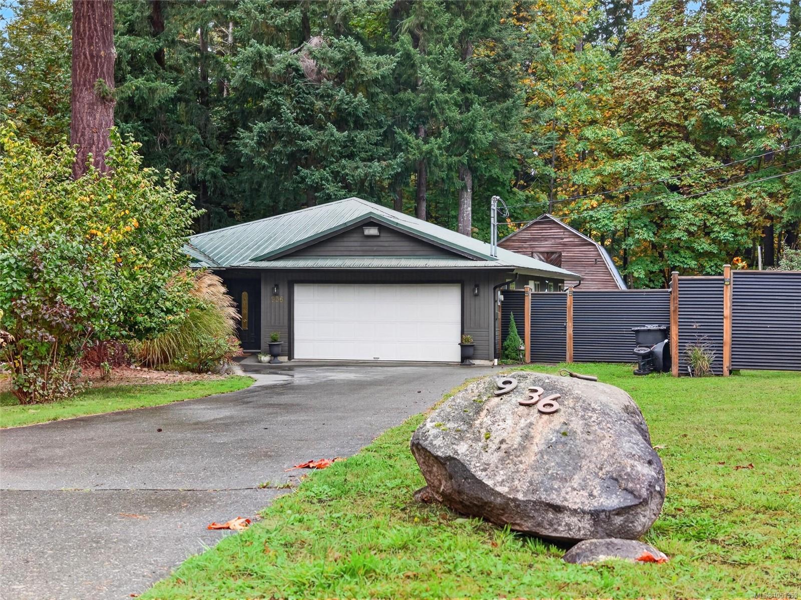 Dark gray home with a white double garage, wooded surroundings, and address 936 displayed on a boulder.