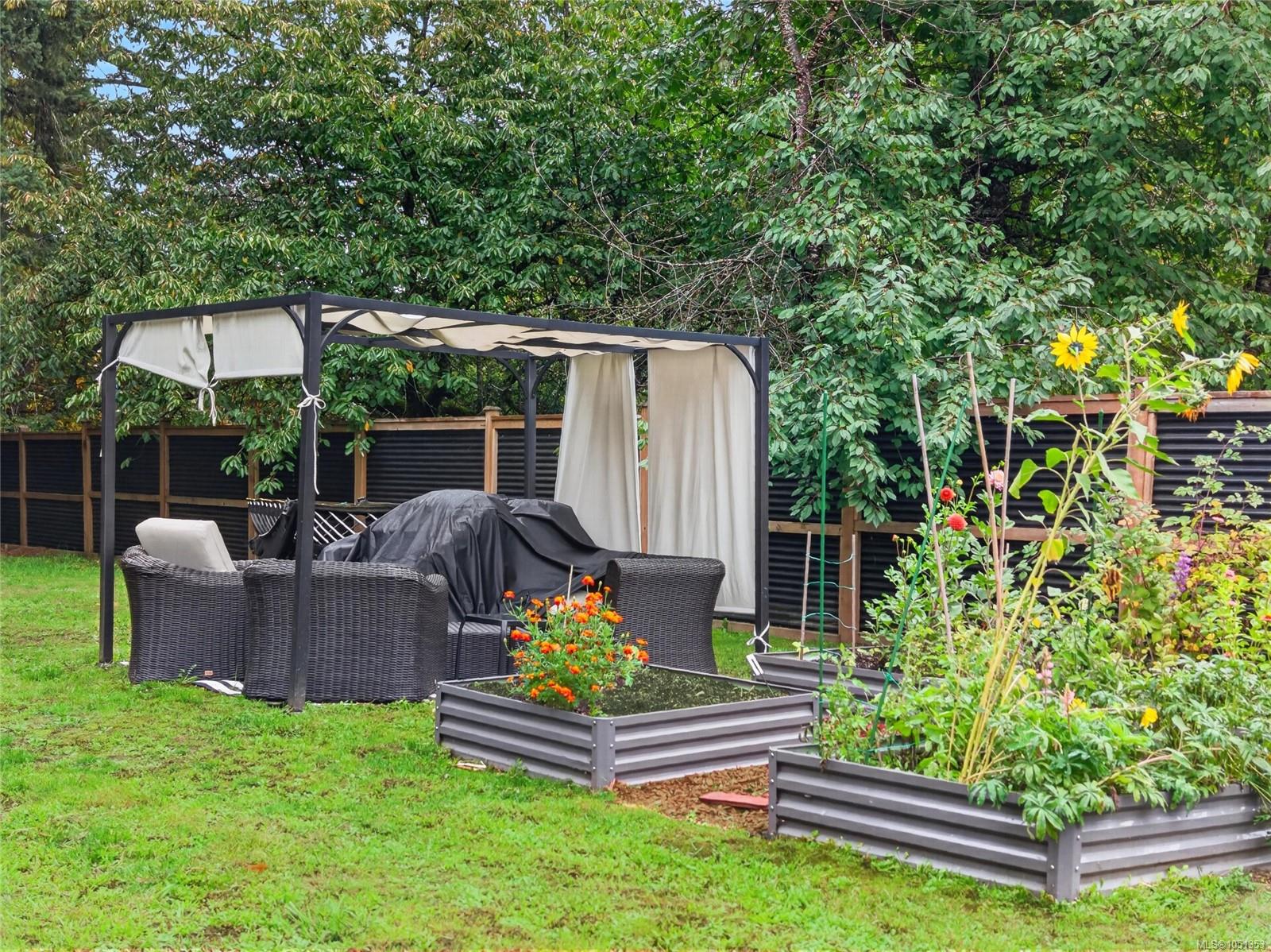 Backyard pergola shelters wicker seating beside raised flower beds and a fenced garden.