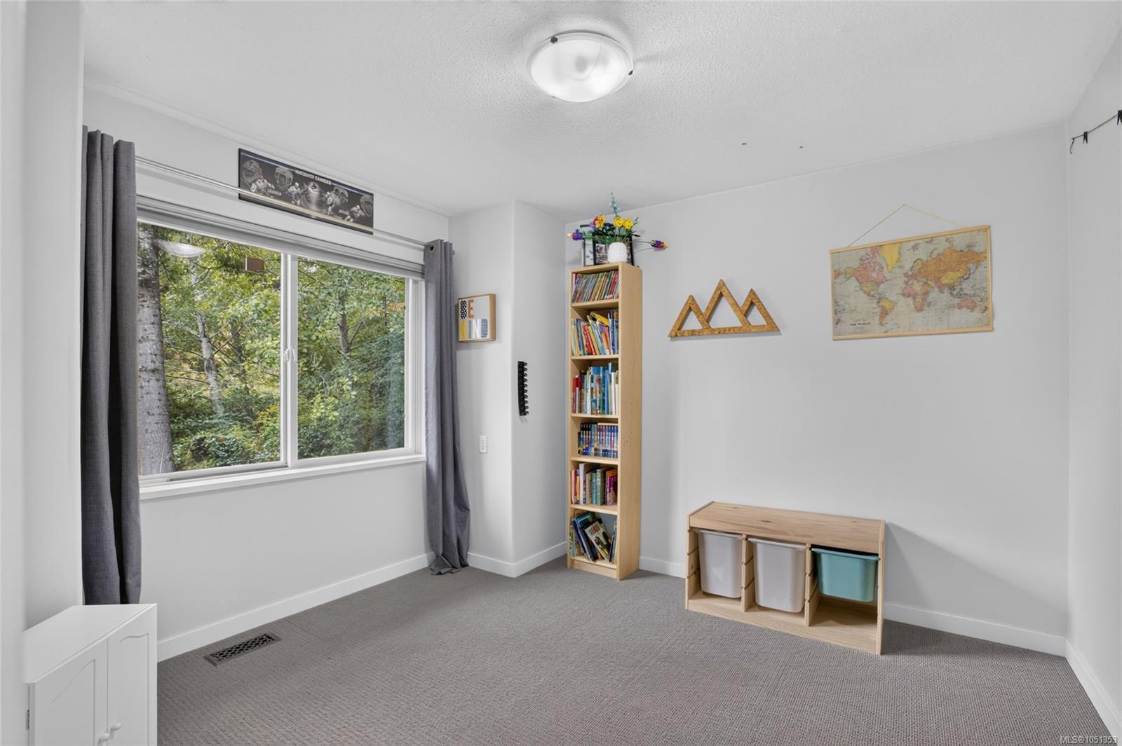 Carpeted bedroom or playroom with a large leafy-window view, bookcase, and toy storage.