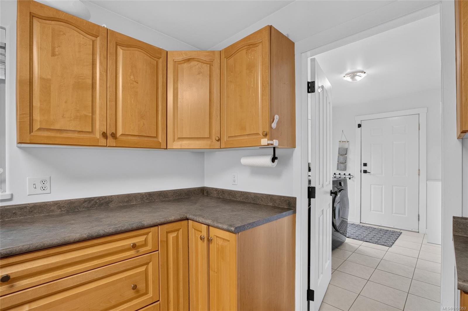 Laundry area with wood cabinets and a gray counter beside a tiled entry and washing machine.