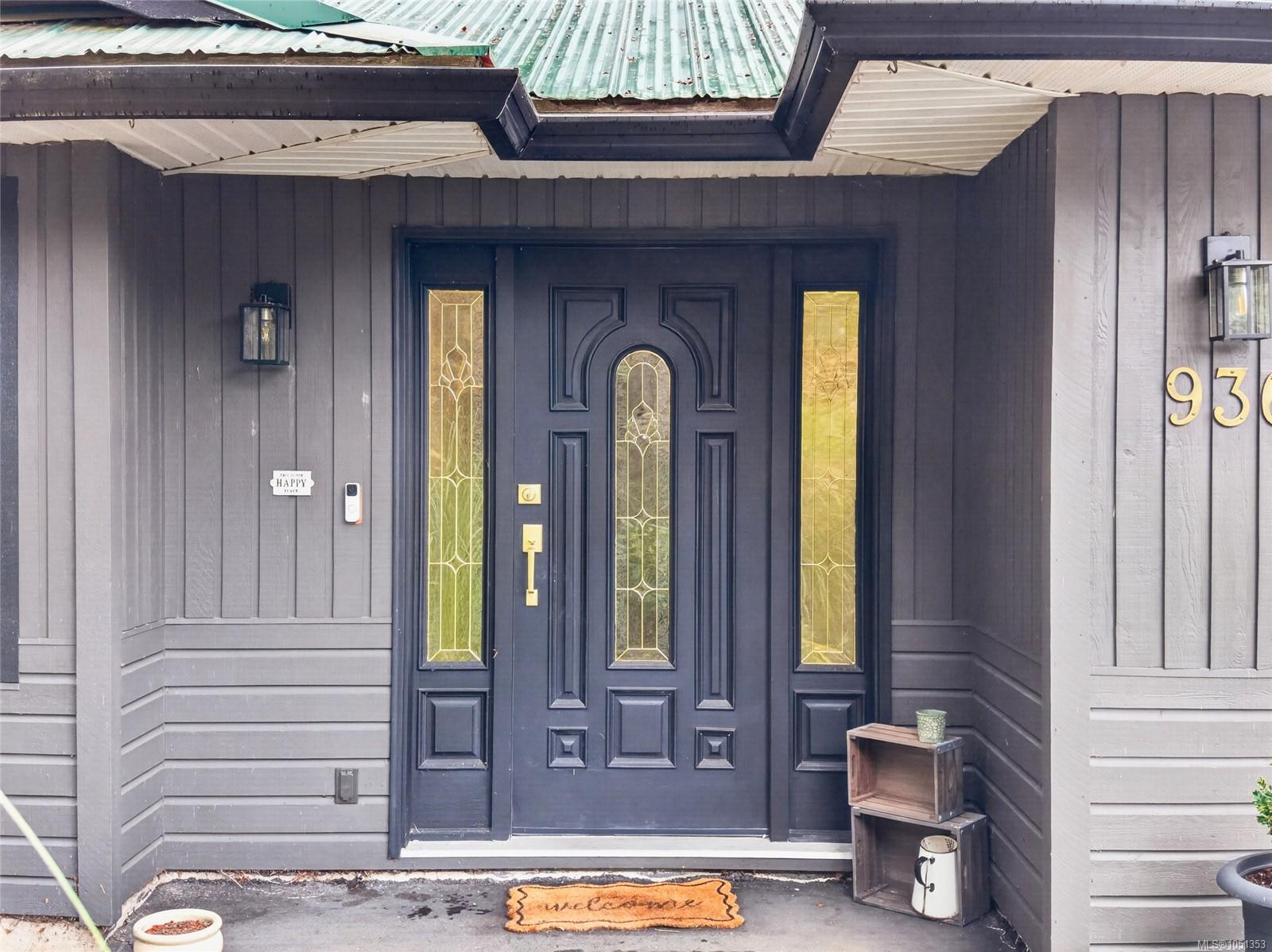 Covered home entrance with a dark decorative door, gold-toned glass panels, and a welcome mat.
