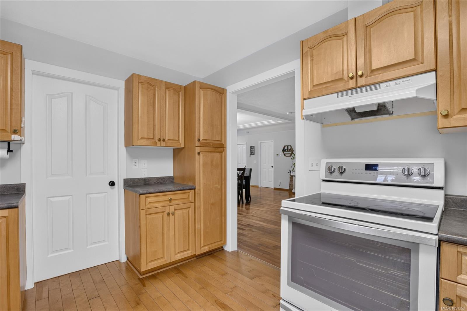 Kitchen with honey-toned cabinets, wood flooring, and a white electric range beside an open doorway.