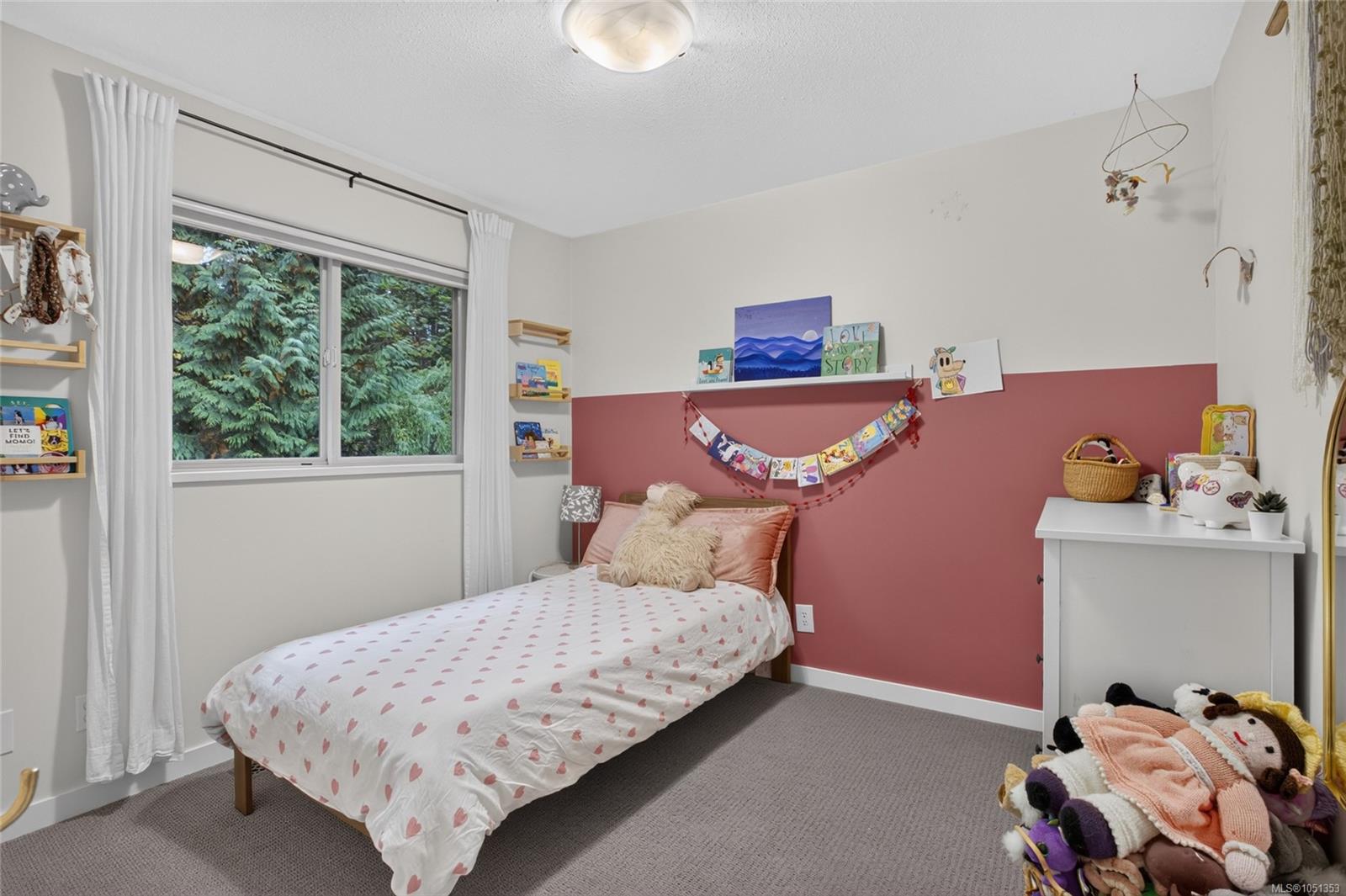 Child’s bedroom with a single bed, dusty rose accent wall, toys, and a window facing evergreen trees.