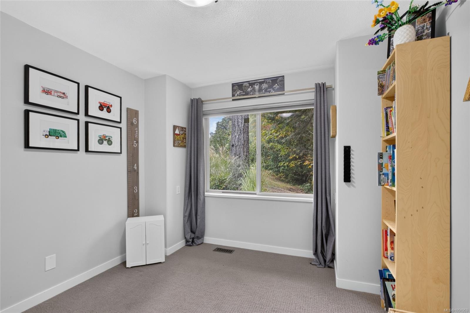Carpeted room with a wide window, gray curtains, framed vehicle prints, and a tall bookcase.
