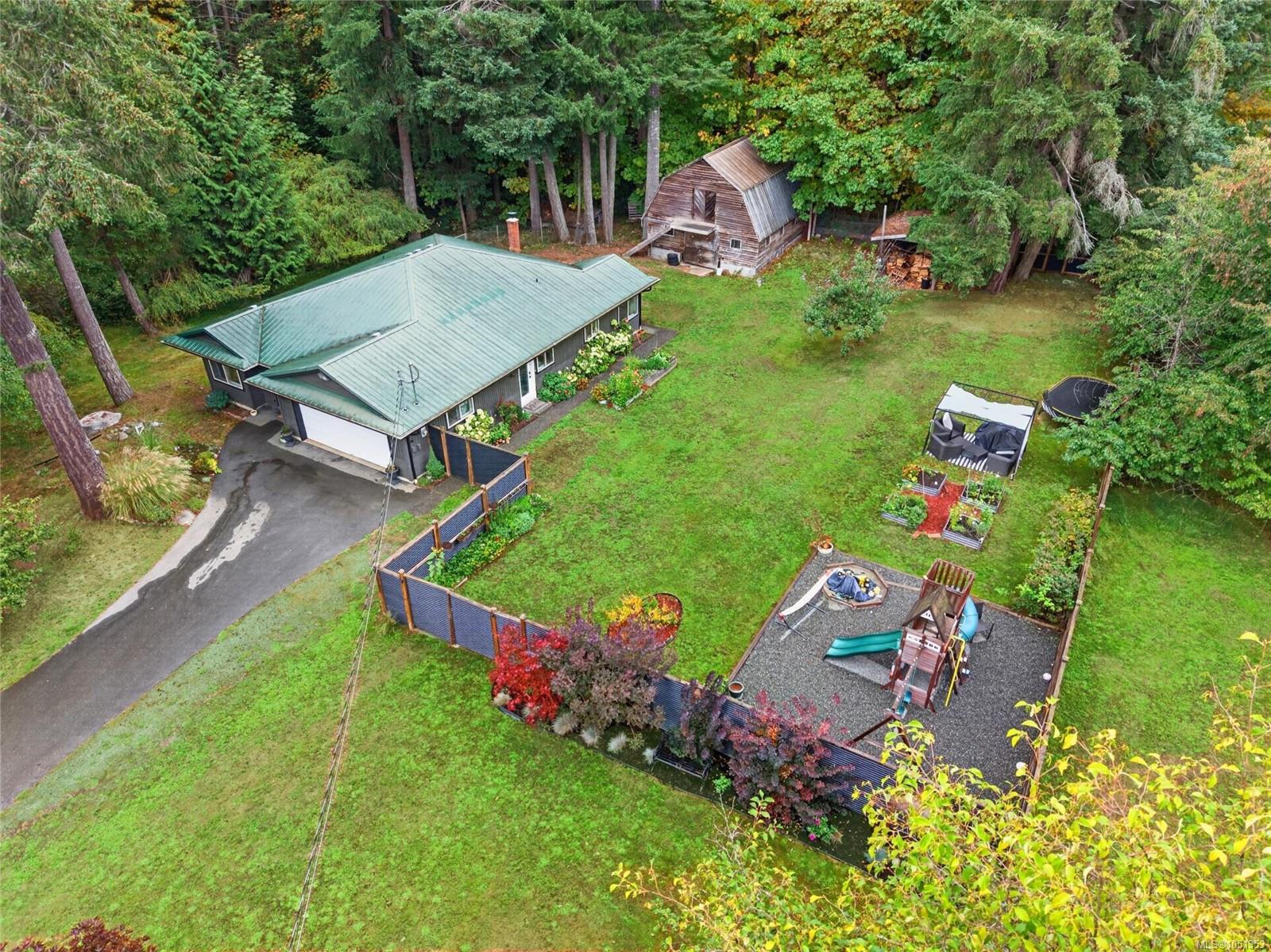 Aerial view of a home, grassy yard, wooden barn, garden beds, and fenced play area among mature trees.