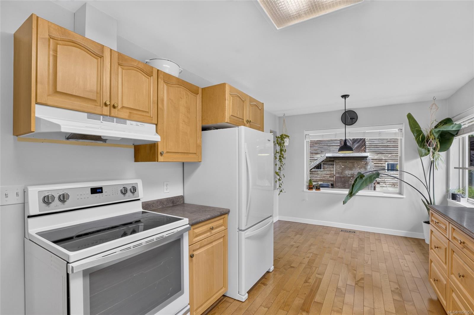 Kitchen with honey-colored cabinets, white appliances, hardwood floors, and a large leafy plant by the windows.