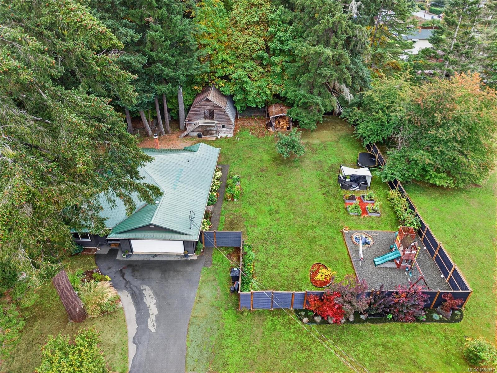 Aerial view of a tree-surrounded home with a large lawn, fenced playground, garden beds, and wooden outbuilding.
