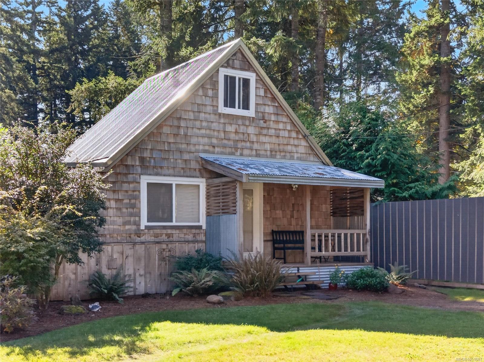Cedar-shingle cottage with a steep roof and covered porch, nestled among trees beside a privacy fence.