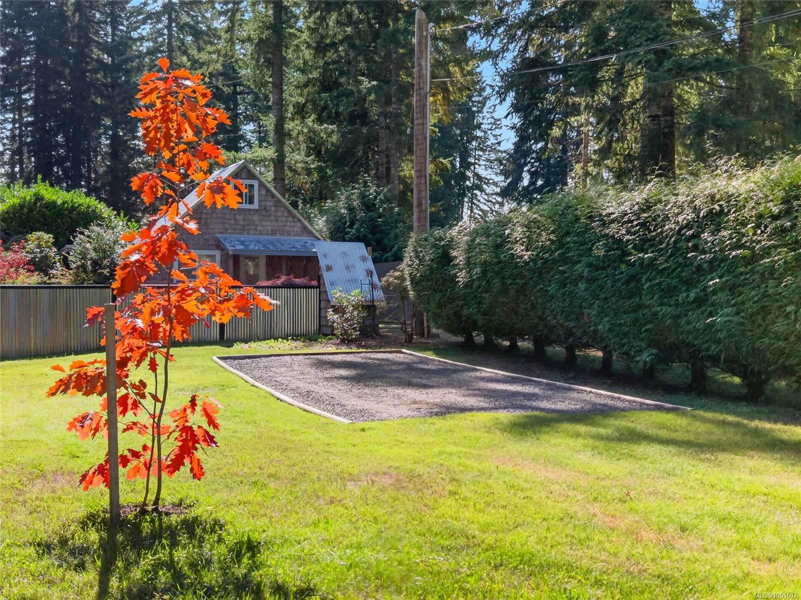 Landscaped yard with a gravel court, small shingled house, tall hedge, and orange-leaved tree.