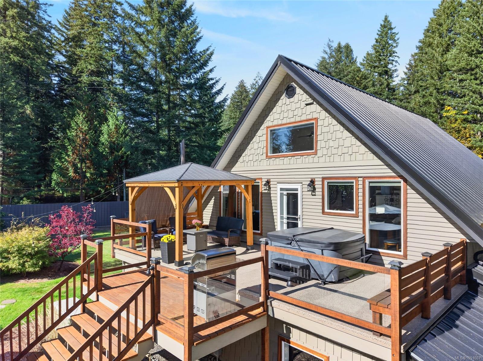 Gabled house with a raised deck, covered seating area, and hot tub among evergreen trees.