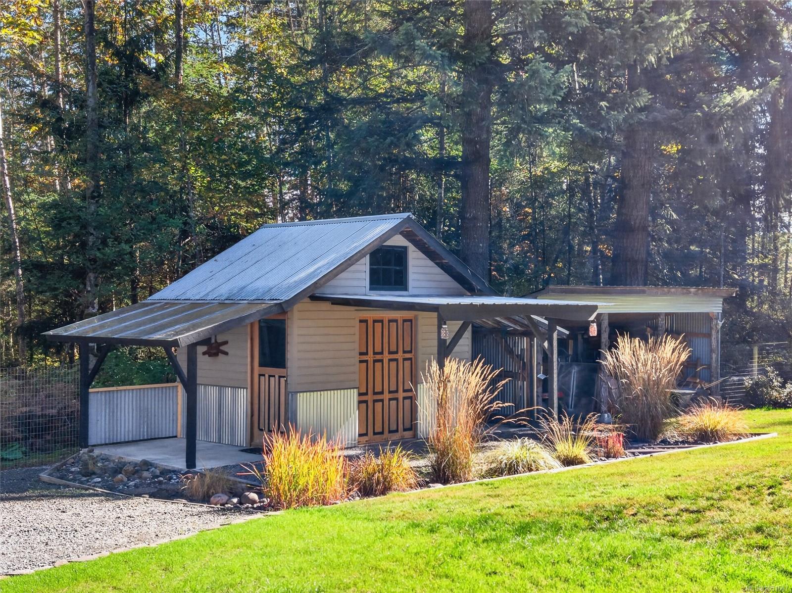 Wood-sided building with a metal roof and covered parking, set beside a gravel drive among trees.