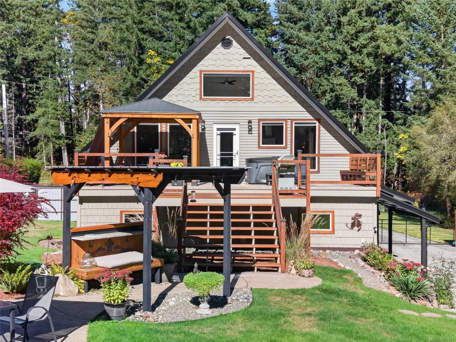 Gray A-frame house with a raised wood deck, broad stairs, and covered patio in a wooded garden.