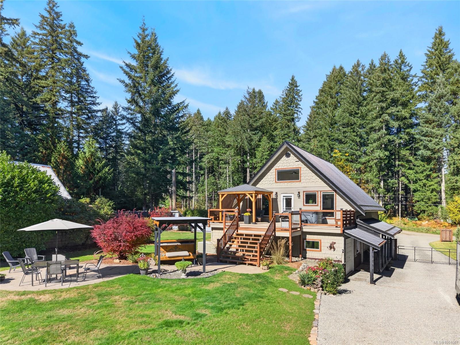 Beige A-frame house with a raised wooden deck, landscaped lawn, and gravel driveway among evergreen trees.