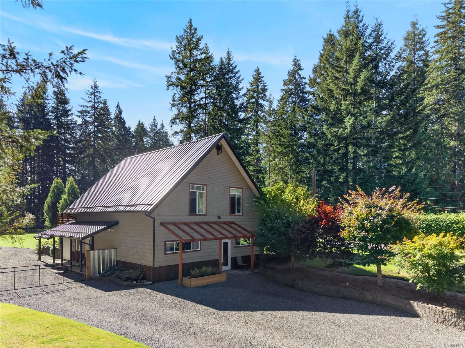 Gray home with a steep metal roof and timber pergola, surrounded by evergreens and gravel driveway.