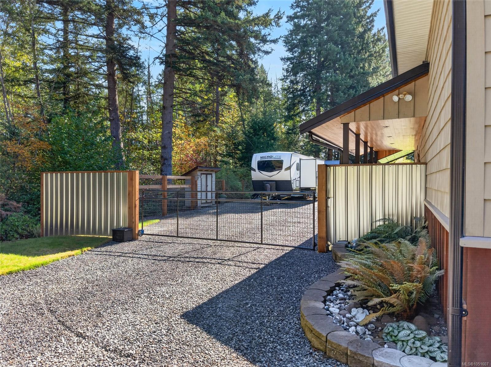 Gated gravel driveway beside a house, with a travel trailer and forested surroundings.