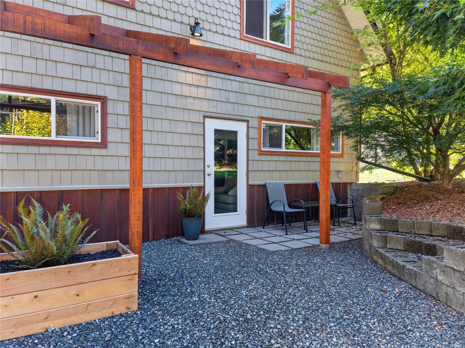 House courtyard with a timber pergola, gravel paving, patio chairs, and a raised fern planter.