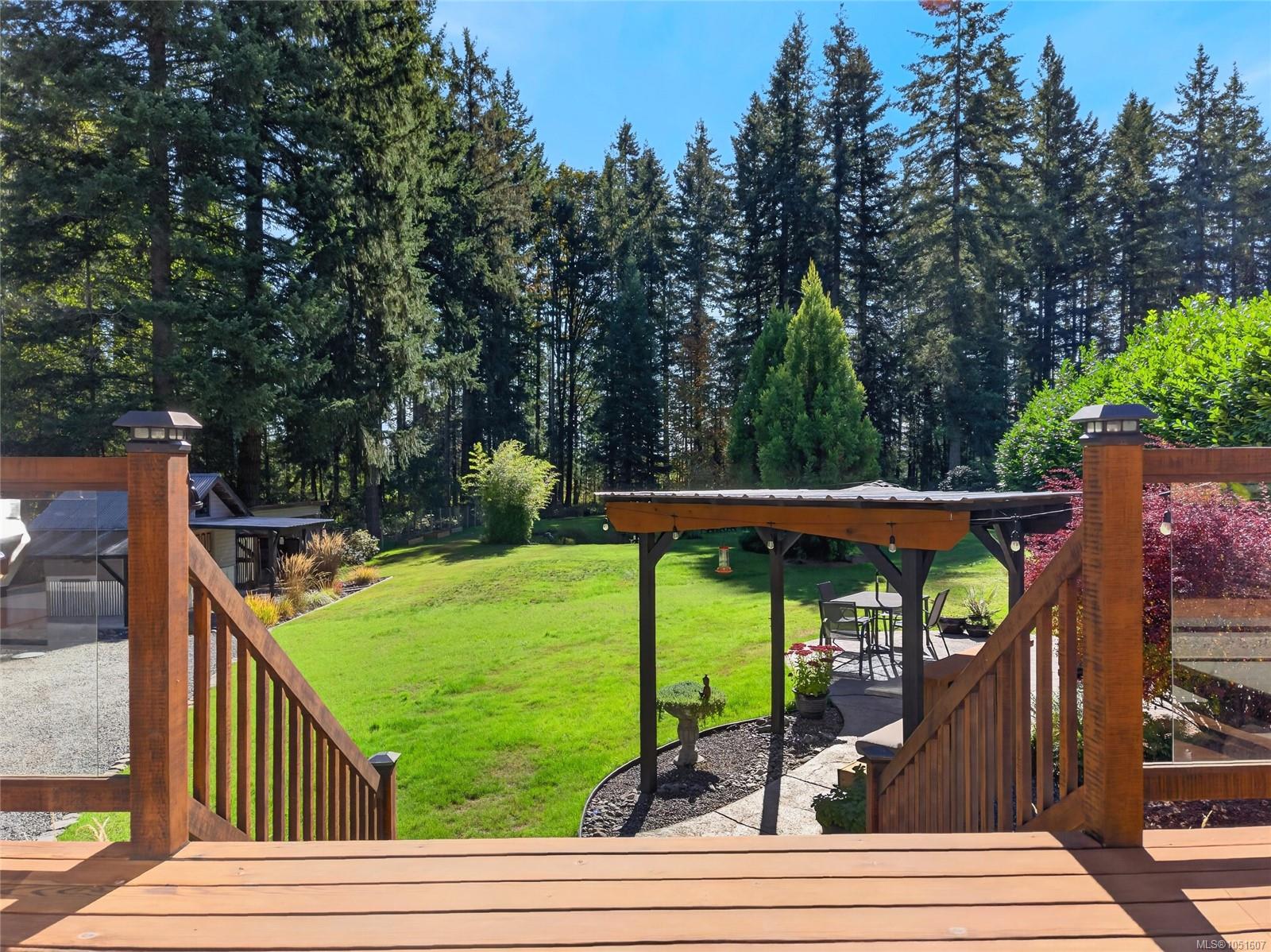 Wooden deck overlooks a green lawn, pergola-covered patio, and tall evergreen trees.