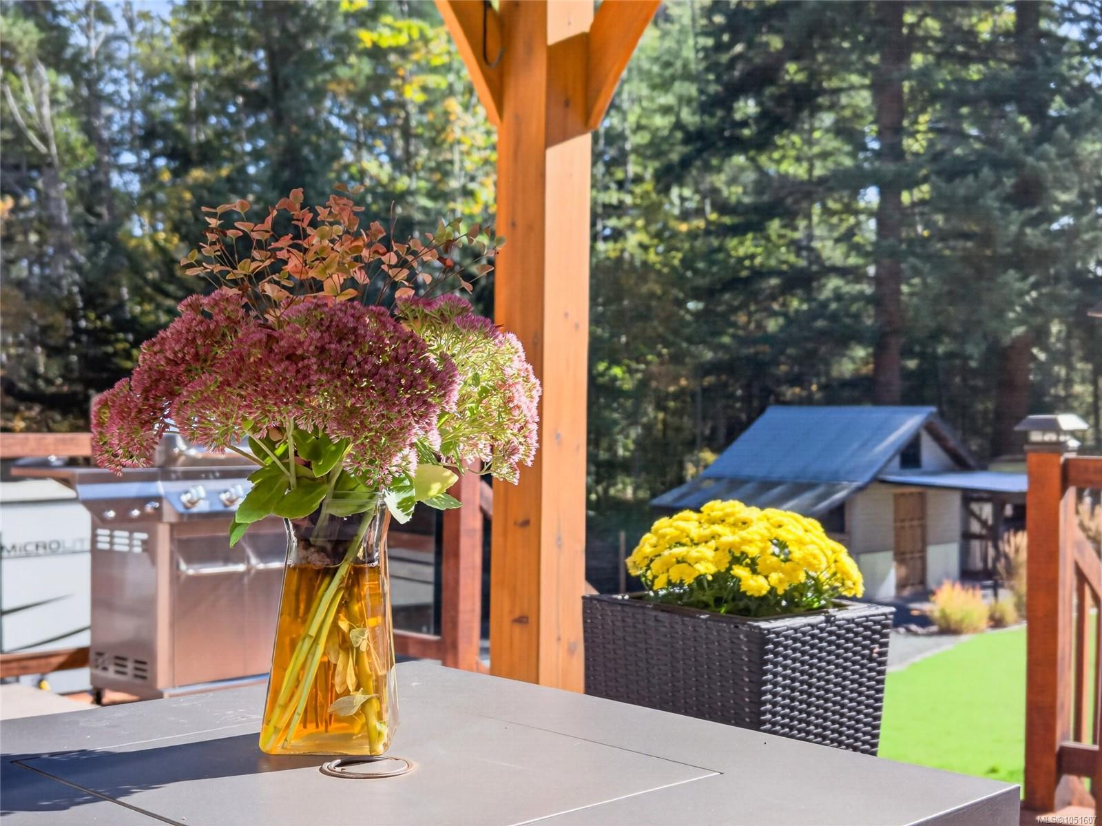 Outdoor deck with pink flowers on a table, a barbecue, and a yellow-flower planter overlooking trees.