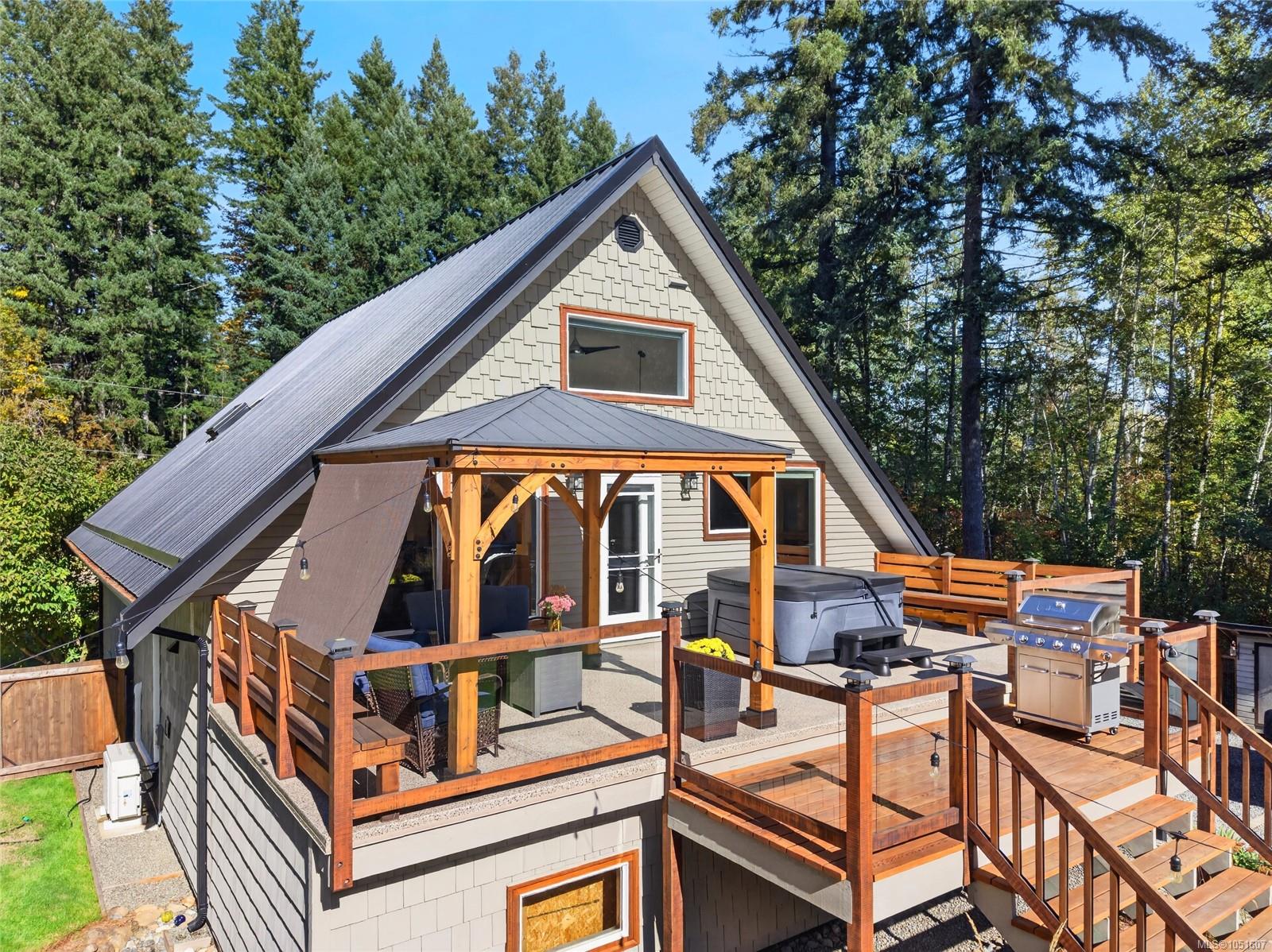 Gray A-frame house with a covered deck, hot tub, and grill among mature trees.