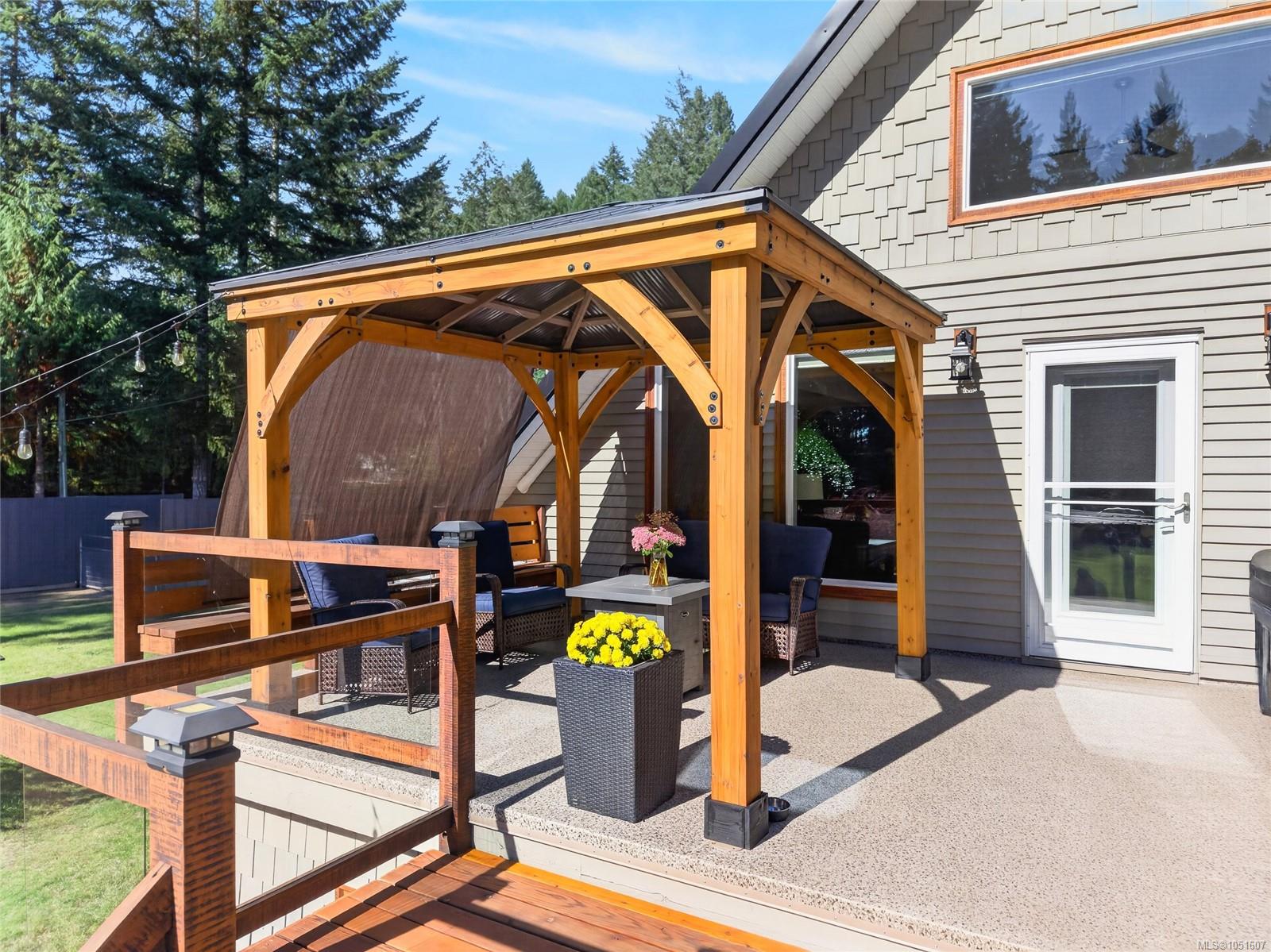 Raised deck with a timber pergola, glass railing, outdoor seating, and potted flowers beside a gray house.