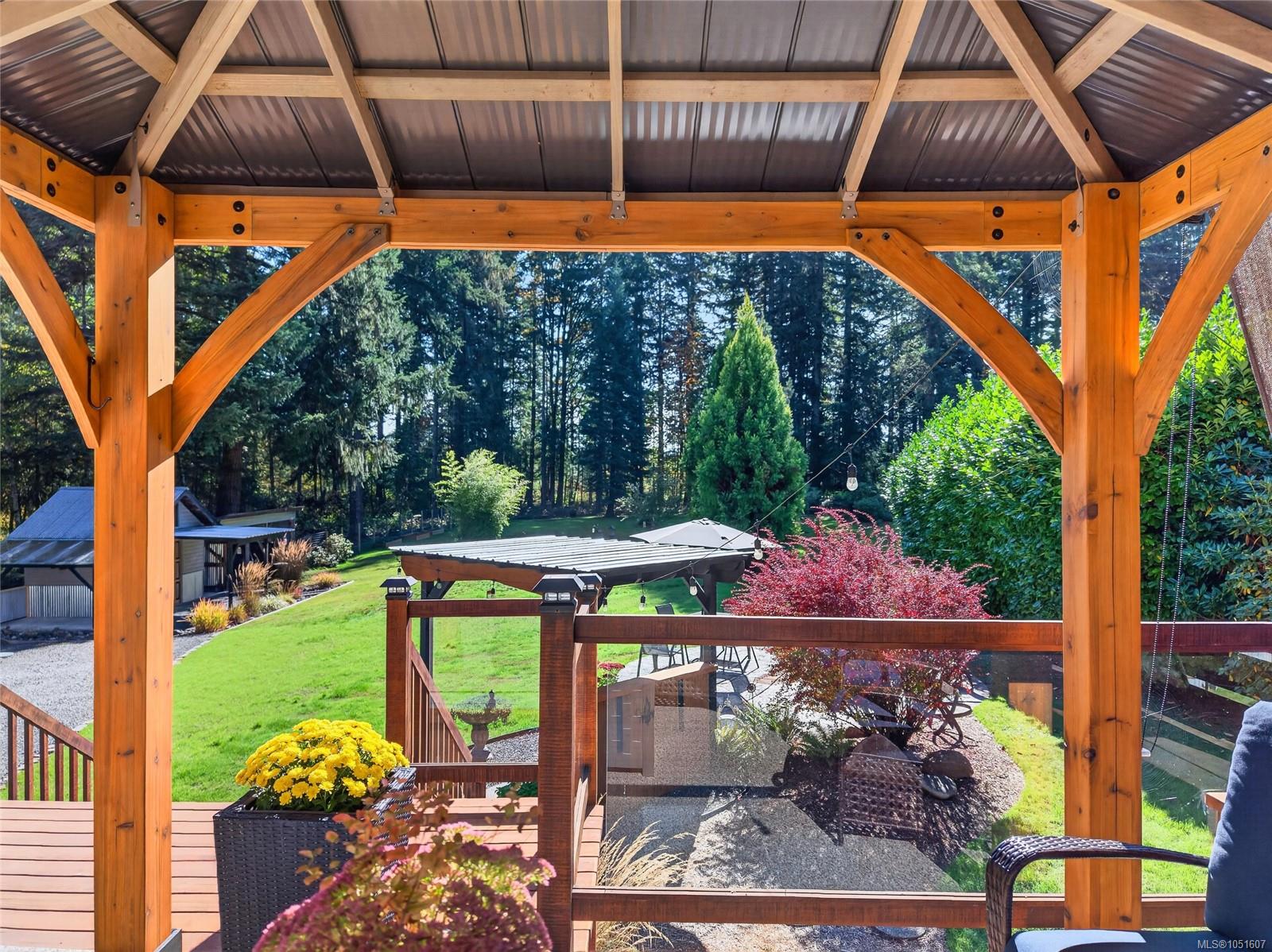 Covered timber deck overlooking a landscaped backyard with lawn, gravel patio, flowers, and mature trees.
