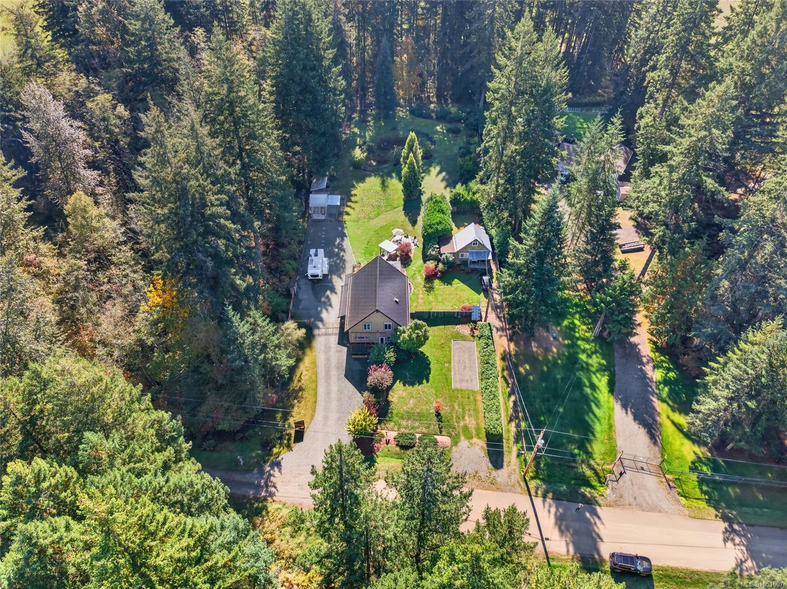 Aerial view of a forest-surrounded home with a long driveway, lawn, outbuildings, and garden beds.