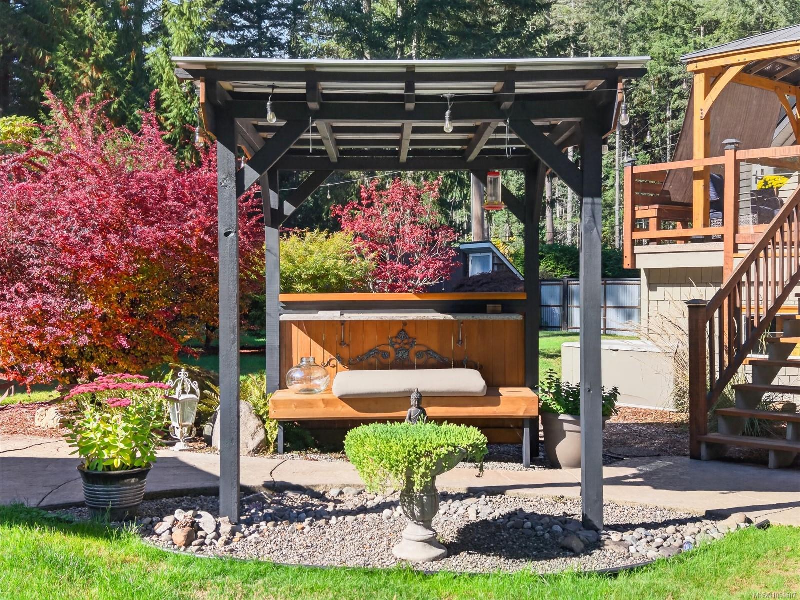 Landscaped backyard with a covered pergola, wooden built-in bench, and raised deck among red-leaved trees.