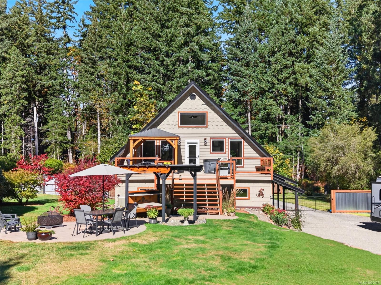 Gray A-frame house with a raised deck, patio seating, and lawn, surrounded by tall trees.