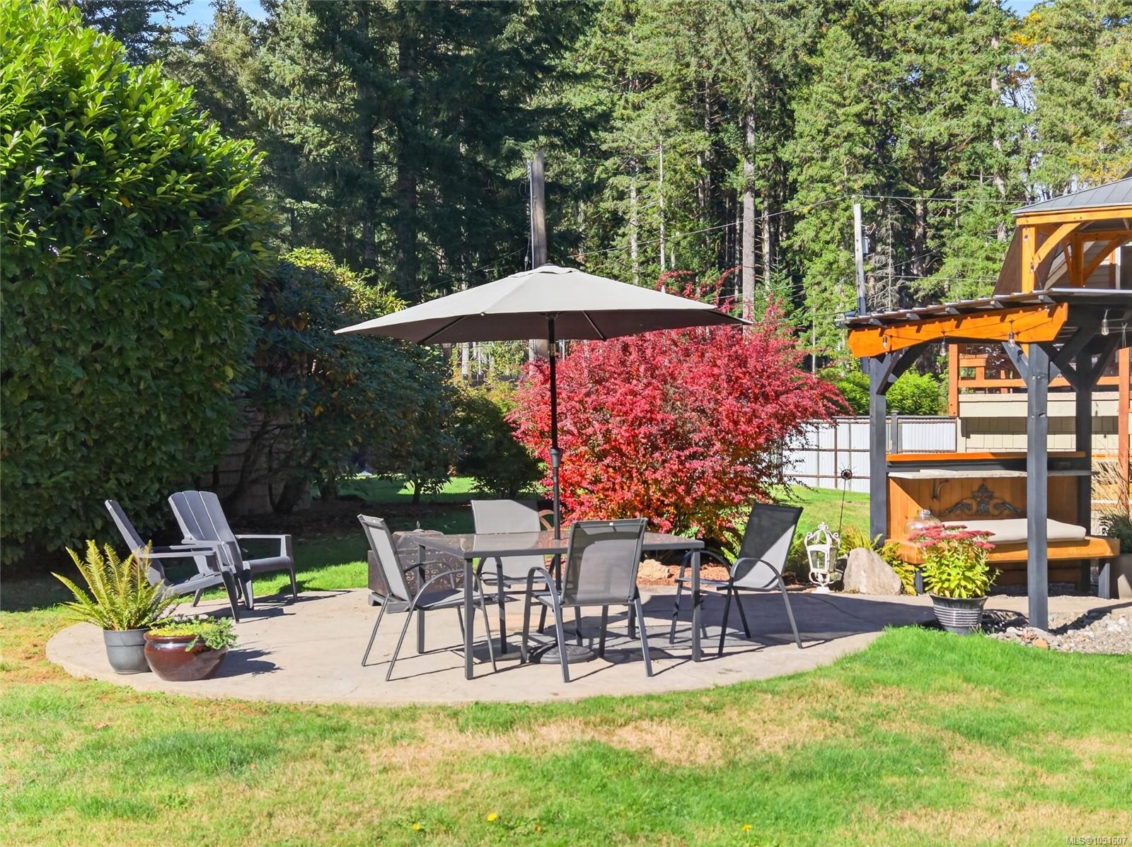 Backyard patio with umbrella-shaded dining set, timber outdoor kitchen, and red-leaved trees.