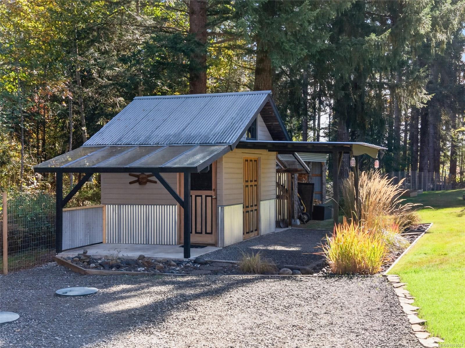Small wooden outbuilding with a steep metal roof in a tree-lined clearing beside a lawn.