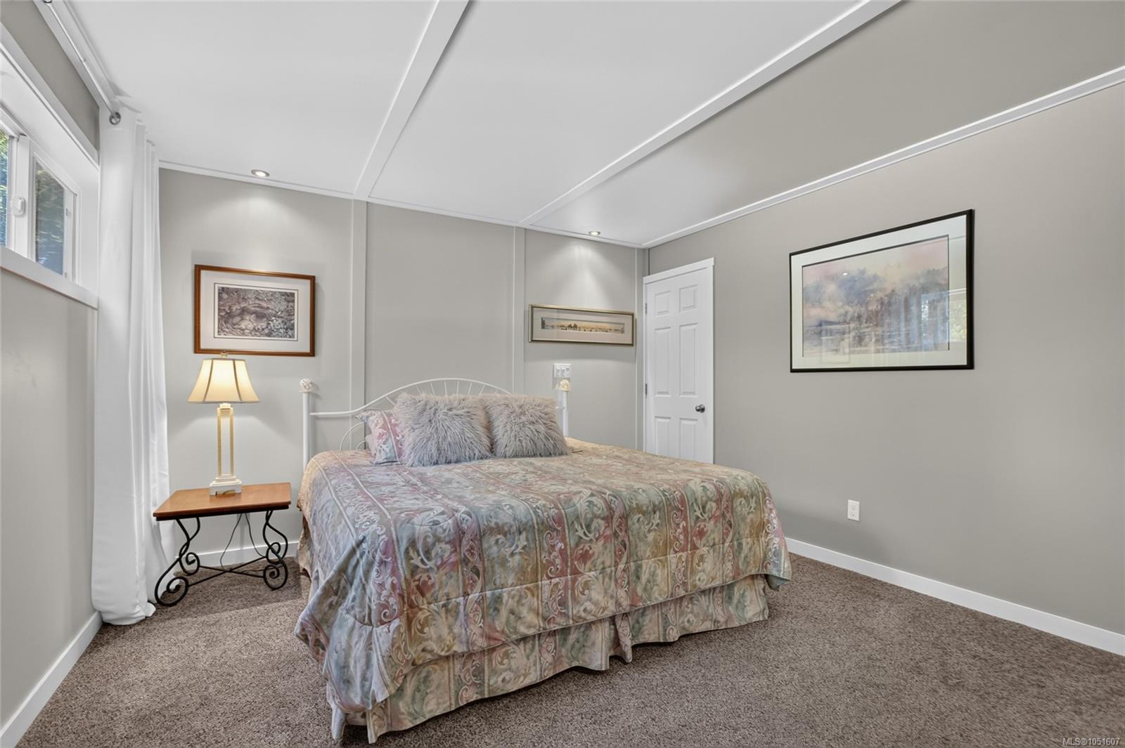 Furnished bedroom with a patterned bedspread, gray walls, framed artwork, and a curtained window.