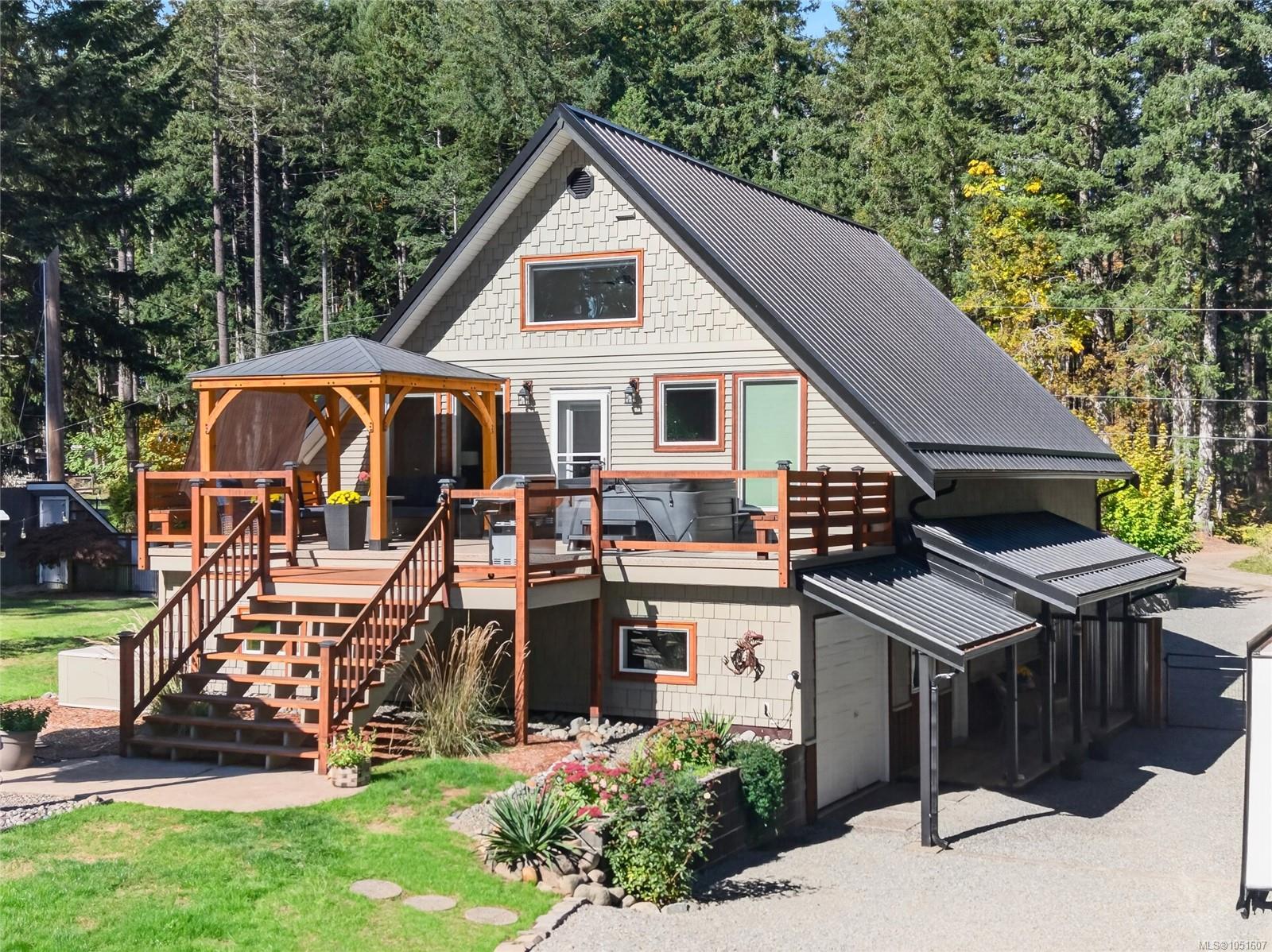Gray A-frame home with a raised wood deck and covered seating area, surrounded by forest.