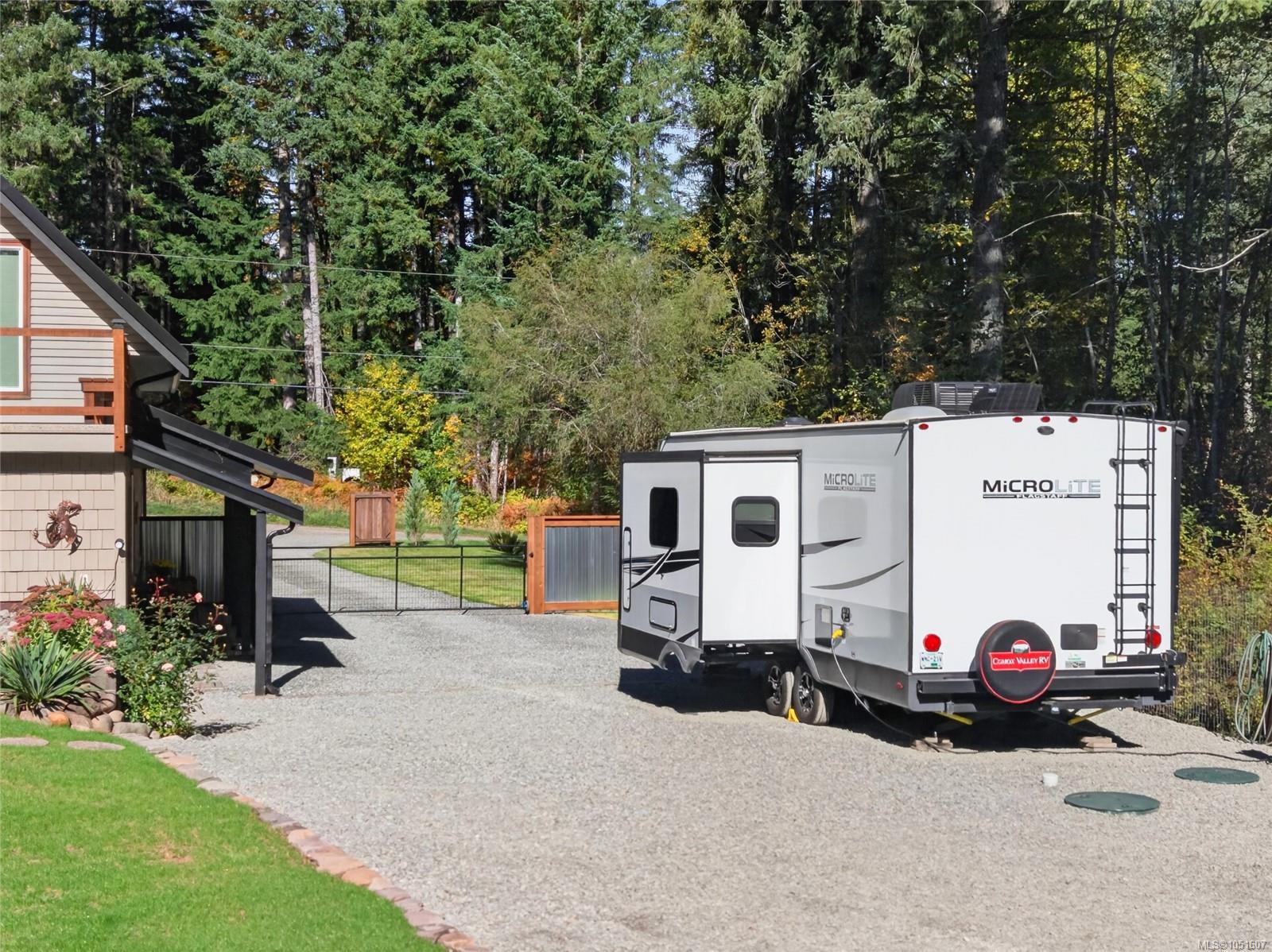 Travel trailer parked on a gravel driveway beside a house, with a wooded yard beyond.