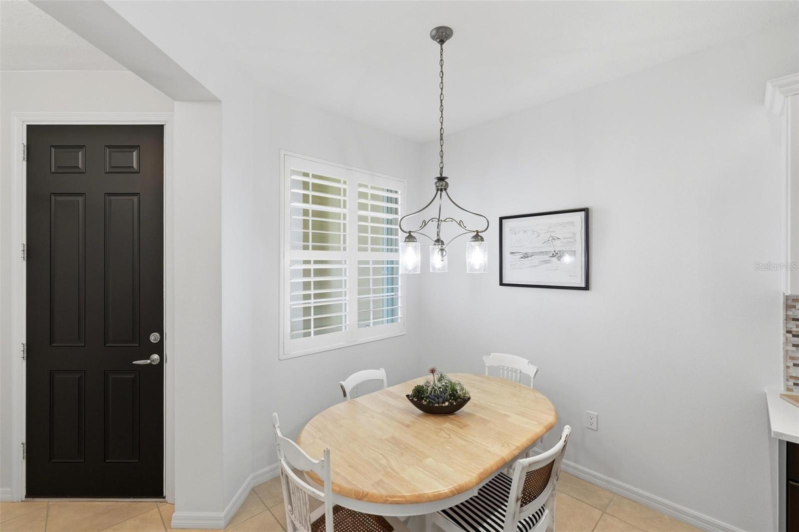Dining nook with a light wood table, white chairs, plantation shutters, and a black front door.