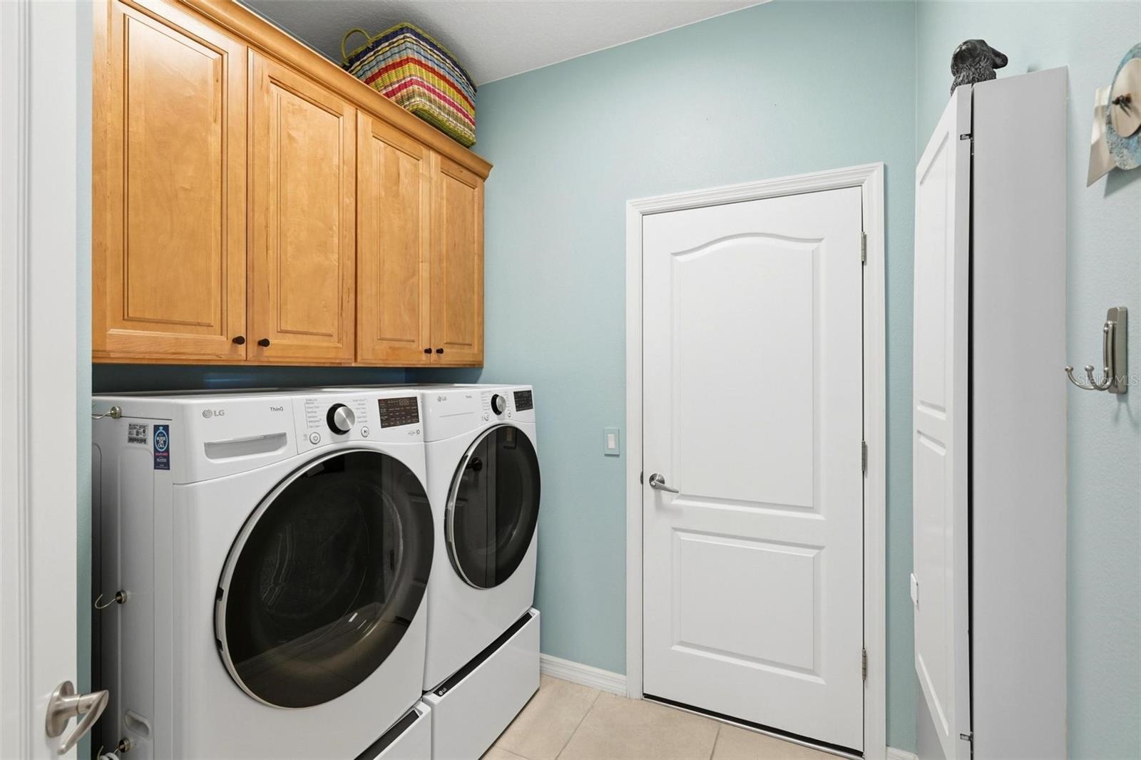 Laundry room with front-load washer and dryer beneath wood cabinets and beside a white storage cabinet.