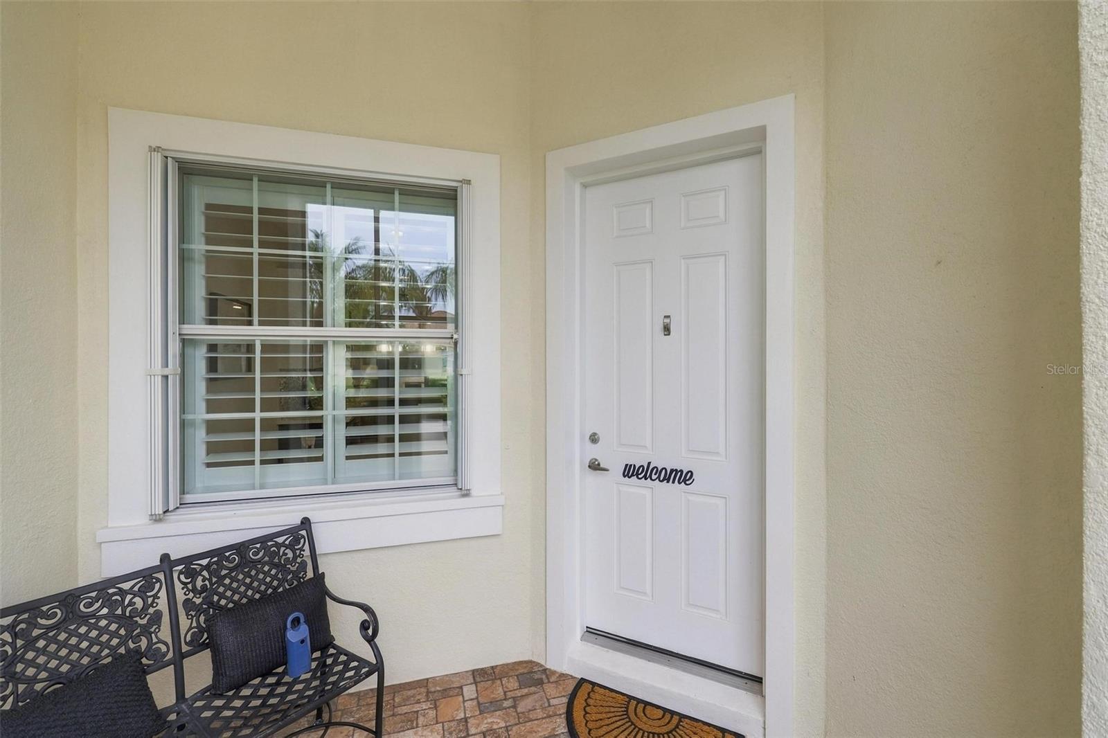 Home entrance with a white front door, window, and black metal bench on a tiled porch.