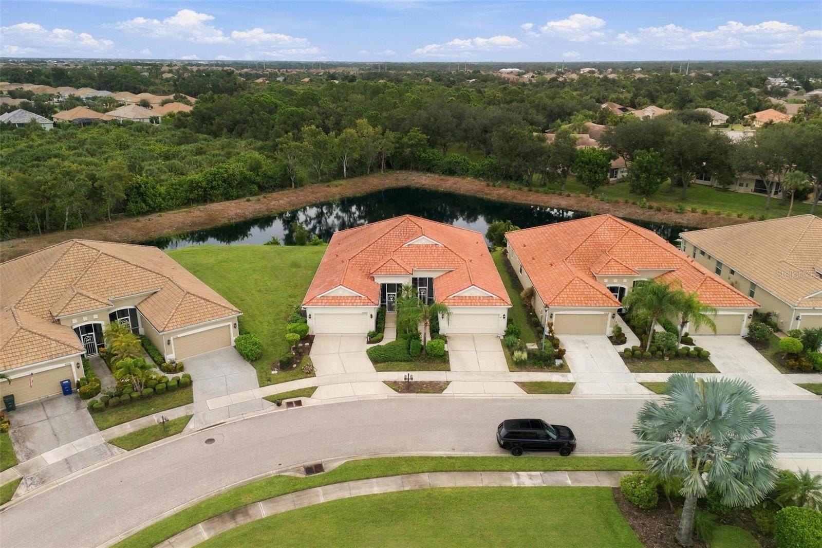 Aerial view of a home beside a pond, with neighboring houses and a tree-lined neighborhood beyond.